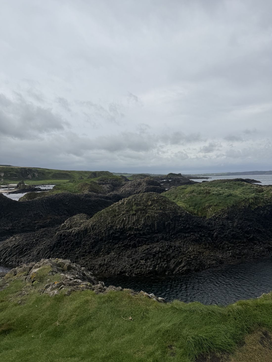 Black basalt rock and green grass on a headland, the sea beyond under a grey sky.