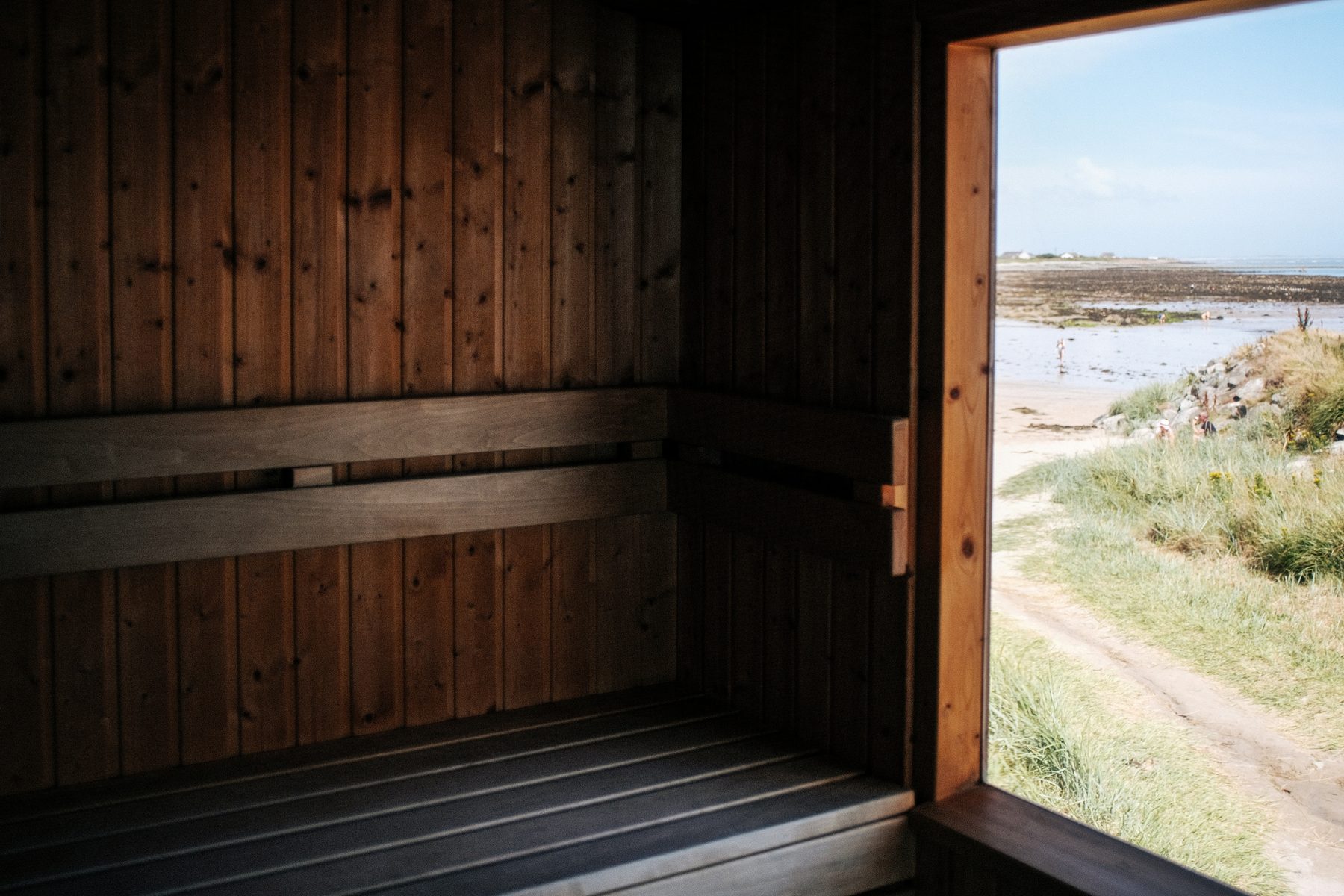 Inside a dark timber sauna, the door open onto dune grass, sand and the sea beyond.