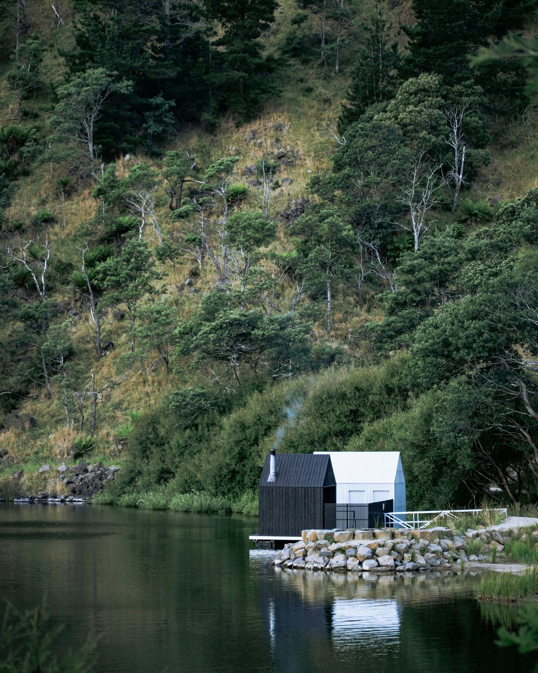 A small dark timber sauna at the edge of a lake, below a steep green hillside.